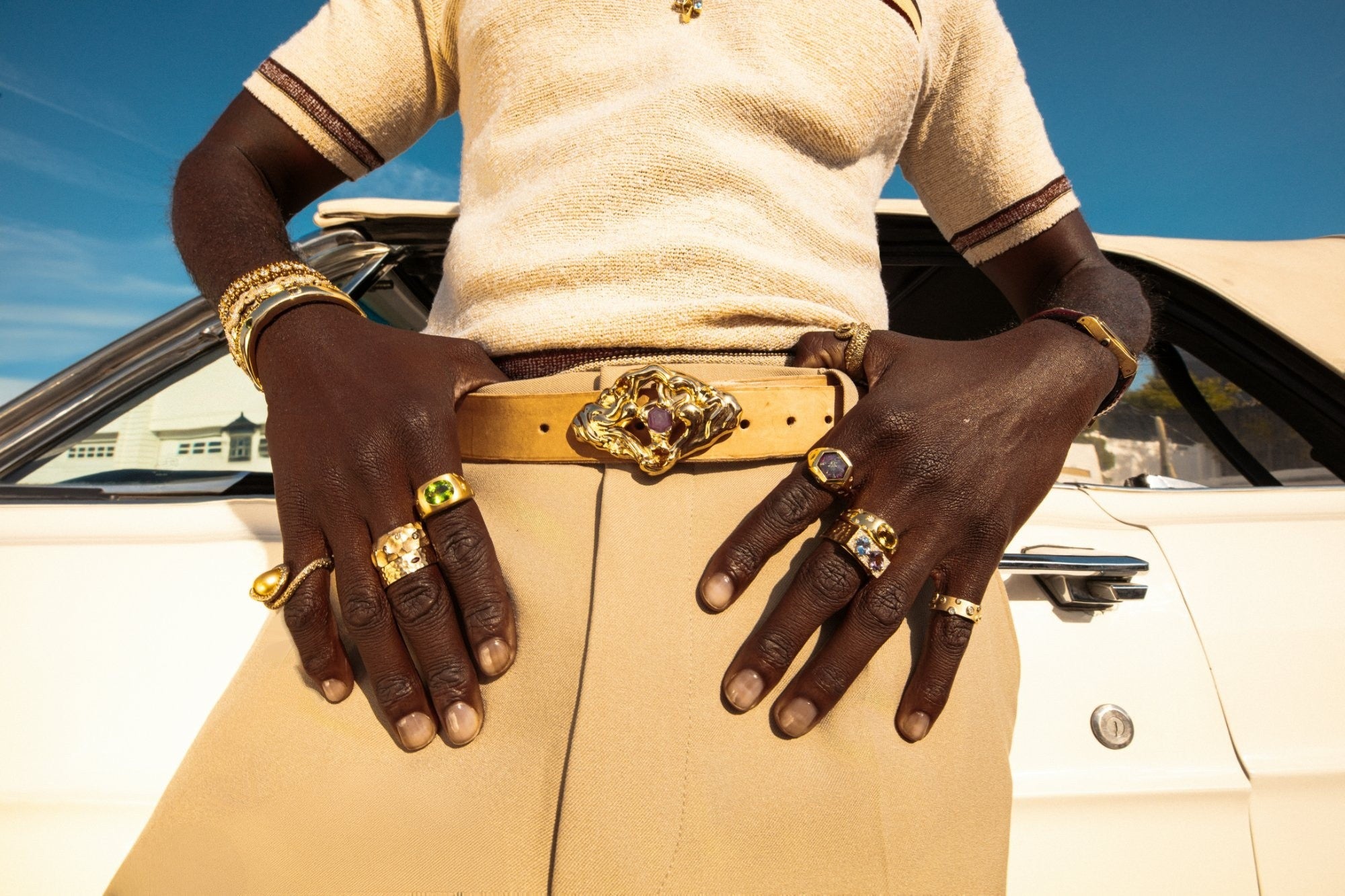 Hands on hips wearing gold gemstone rings and a sculpted gold belt buckle set with a pink sapphire