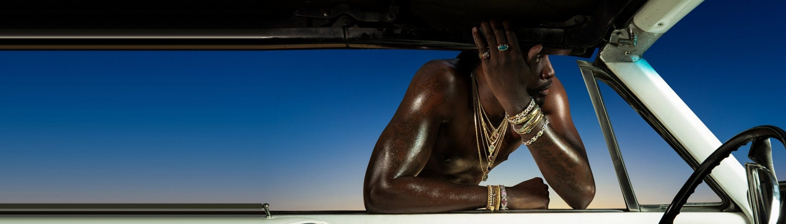 Model leaning through a vintage car window wearing layered gold chains, bracelets and rings